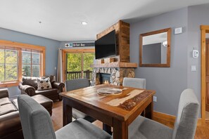 A cozy living room with a stone fireplace, a flat-screen TV mounted above it, and a dining table with chairs. - Short Walk to Lifts & Village Amenities & Private Balcony Overlooks Pool! (Keystone)
