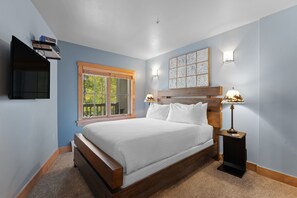 A bedroom with a large bed, a window with blinds, a mounted TV, and two bedside lamps. - Short Walk to Lifts & Village Amenities & Private Balcony Overlooks Pool! (Keystone)