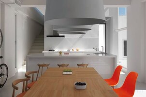 Una cocina moderna con una isla integrada, un comedor con mesa de madera y sillas naranjas, y una escalera que lleva a un nivel superior.