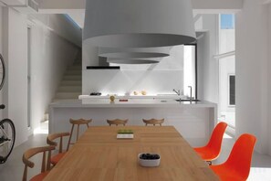 Una cocina moderna con una isla integrada, un comedor con mesa de madera y sillas naranjas, y una escalera que lleva a un nivel superior.