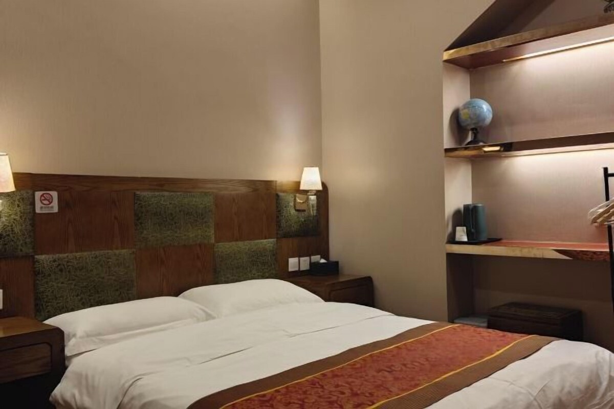 Una habitación de hotel con una cama, mesitas de noche, un estante en la pared con un globo terráqueo y una alfombra roja.