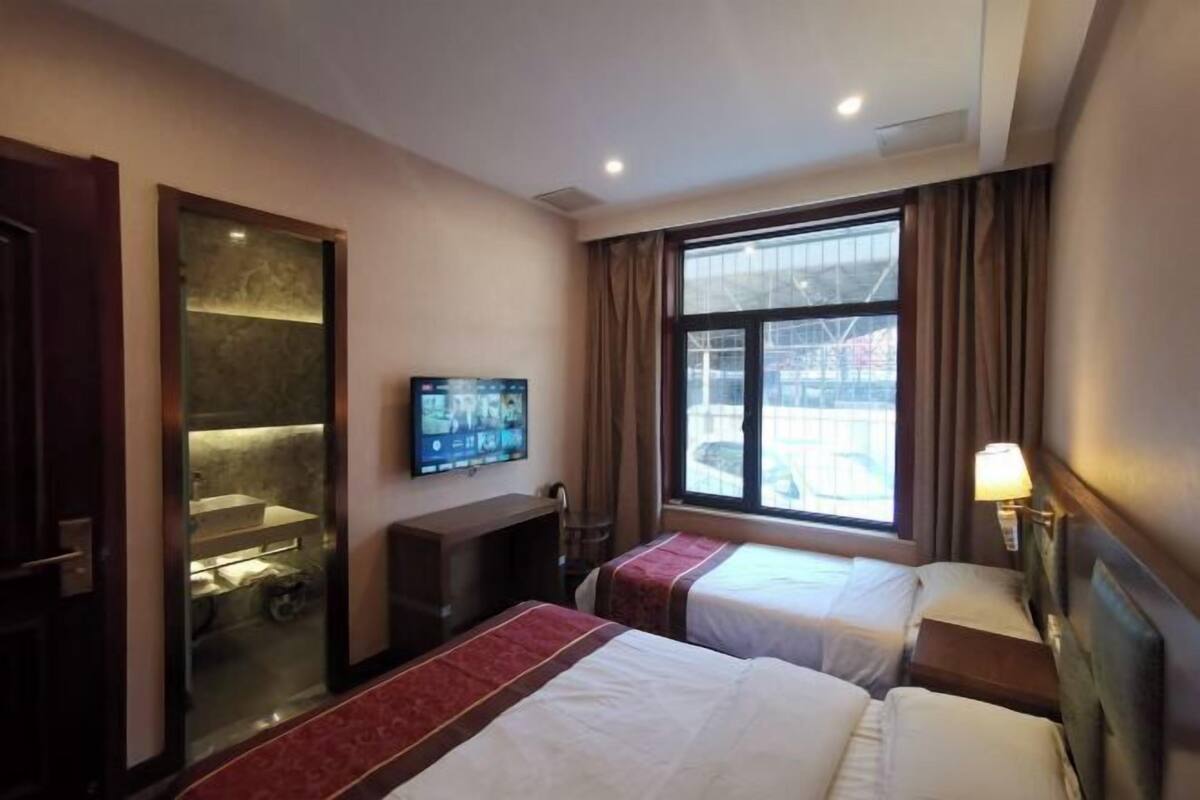 A hotel room with two beds, a TV, a desk, a window with curtains, and a view of the outside.