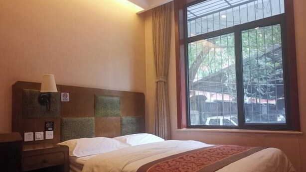 A hotel room with a bed, a nightstand, a window with curtains, and a view of the outside. - Beijing Tiantan Huiqingyuan Hotel (Beijing)