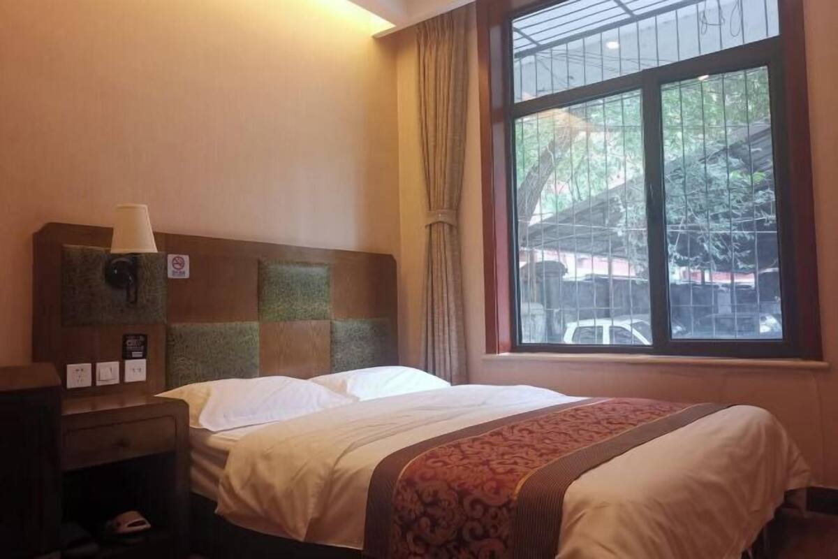 A hotel room with a bed, a nightstand, a window with curtains, and a view of the outside.