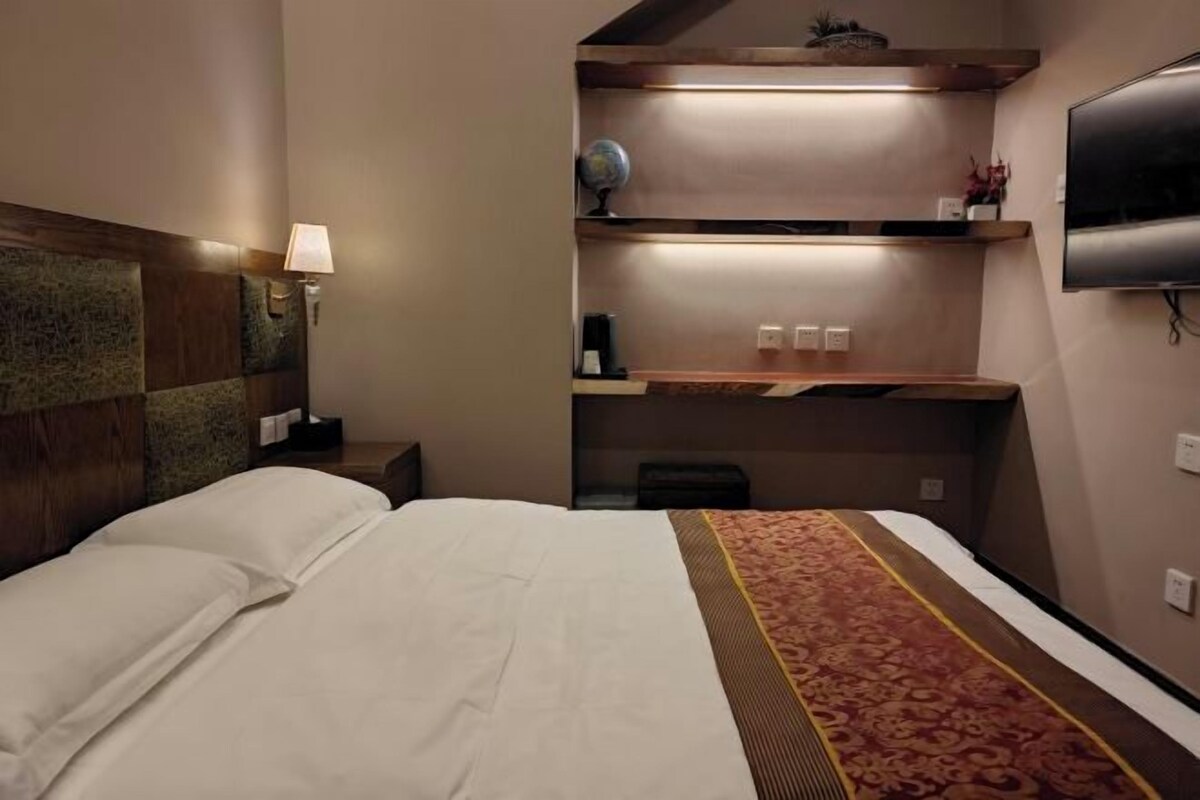Una habitación de hotel con cama, lámpara de noche, estante con adornos y televisor.