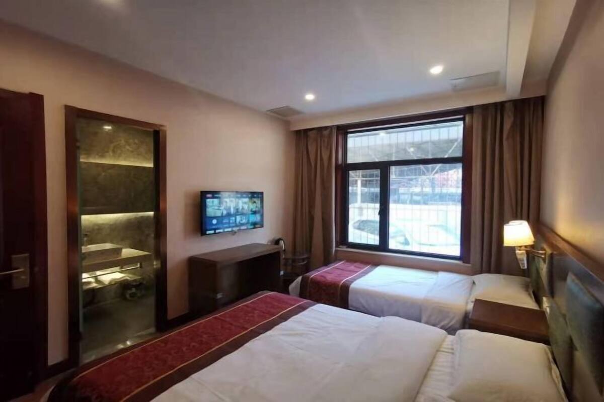 A hotel room with two beds, a television, a desk, a window with curtains, and a shower area.