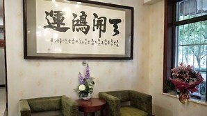 A framed calligraphy piece on the wall, two green armchairs, a small table with a vase of flowers, and a window with a bouquet. - Beijing Tiantan Huiqingyuan Hotel (Beijing)