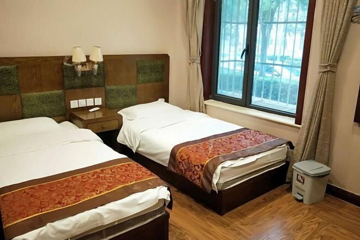 A hotel room with two beds, a nightstand, a window with curtains, and a trash bin.