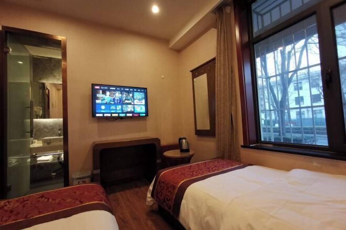A hotel room with two beds, a TV on the wall, a window with a view, and a mirror.