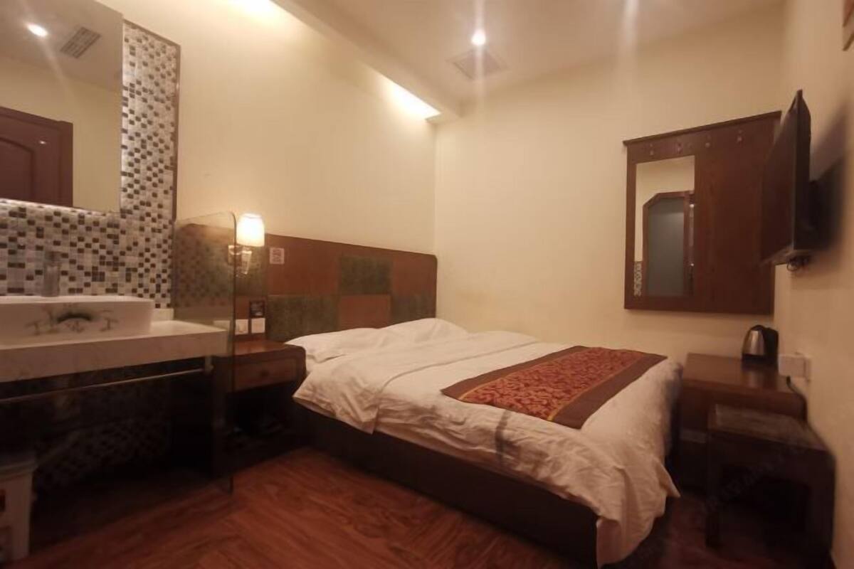 A hotel room with a bed, a bedside table with a lamp, a mirror, and a small table with a kettle.