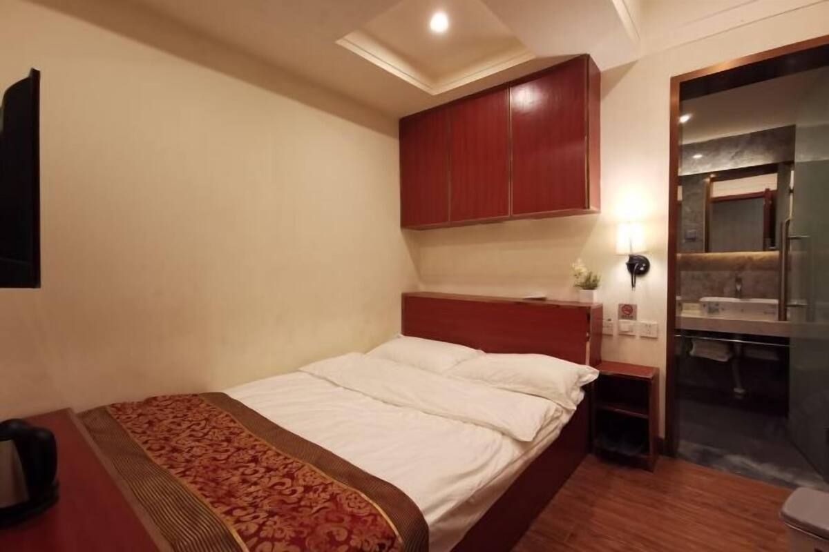 A hotel room with a bed, a wooden headboard, a small bedside table, a nightstand, a mirror, and a bathroom visible through a glass door.