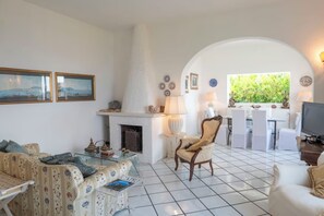A living room with a fireplace, white tiled floor, a sofa, a chair, and a dining area with white chairs and a table.