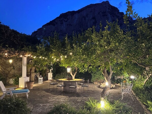 An outdoor patio with stone flooring, a round table, and chairs, surrounded by greenery and a mountain backdrop.