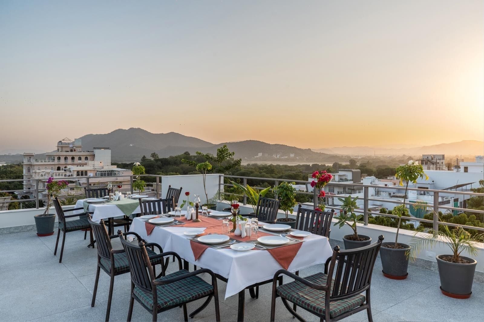 A rooftop dining area with tables set for two, potted plants, and a cityscape view in the background.