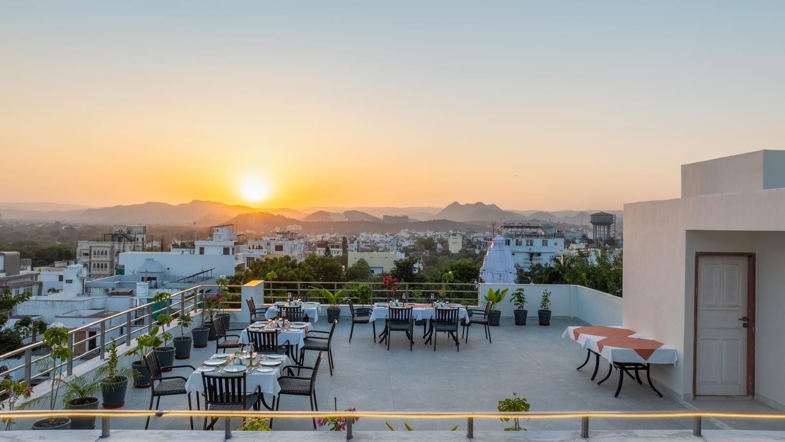 Una terrazza sul tetto con tavoli e sedie, piante in vaso e vista sulla città al tramonto.