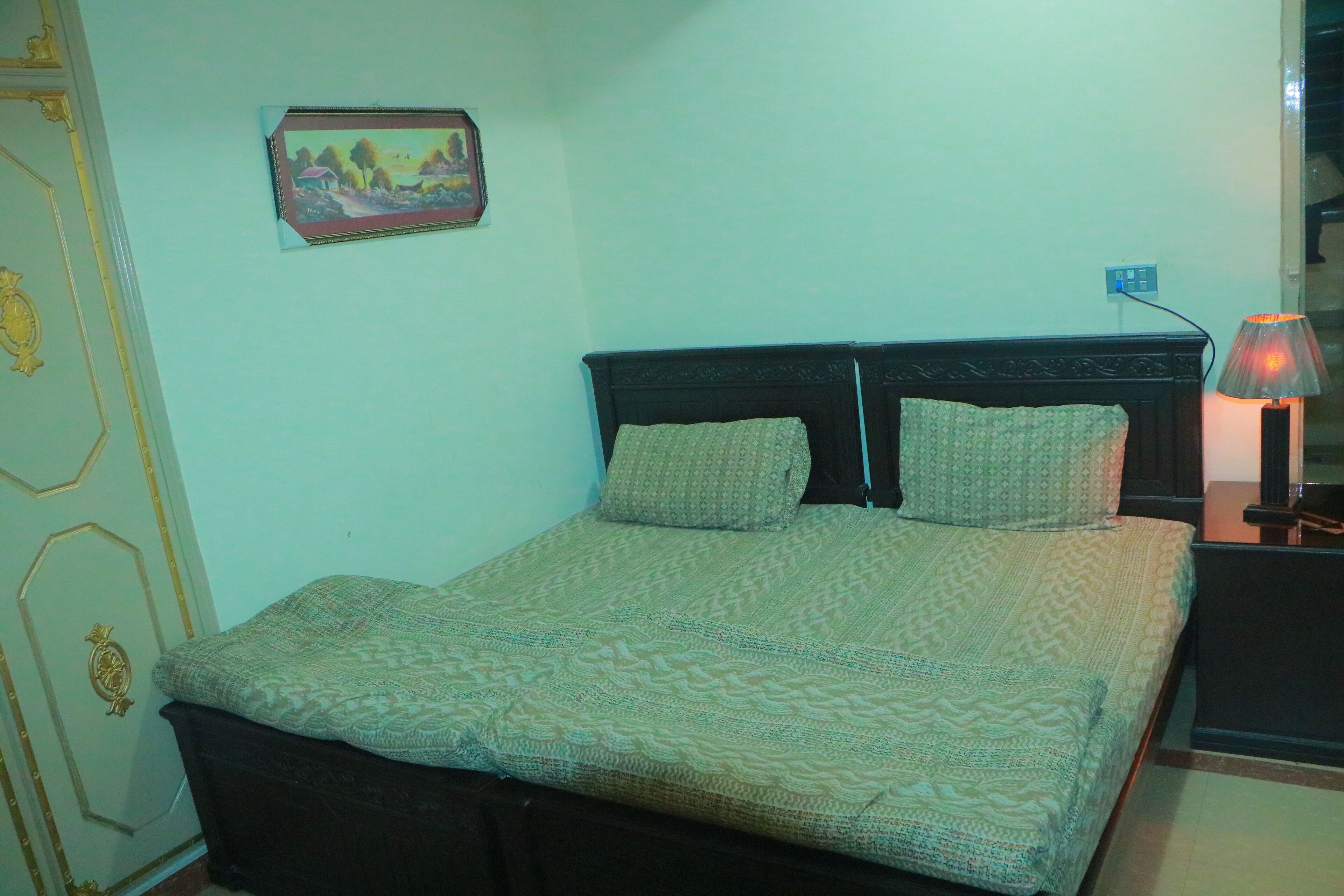 Una cama bien tendida con una colcha estampada y almohadas, una lámpara de noche y un cuadro en la pared.