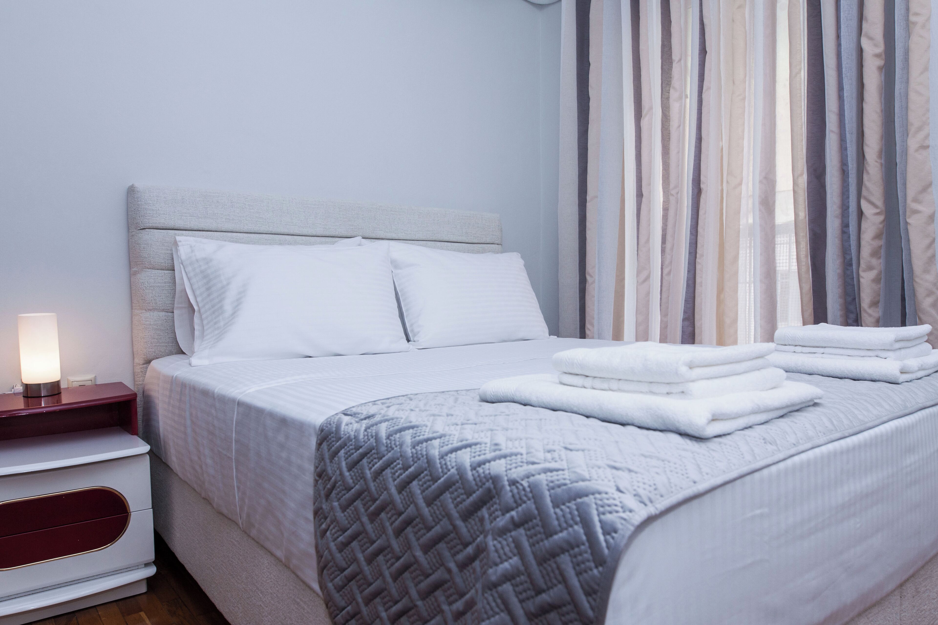 A neatly made bed with white linens, a grey textured blanket, and folded towels on the side.