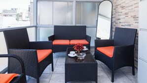 Un balcon agrémenté de meubles en osier, de coussins orange et d’une table avec un plateau en verre sur laquelle est posé un vase.