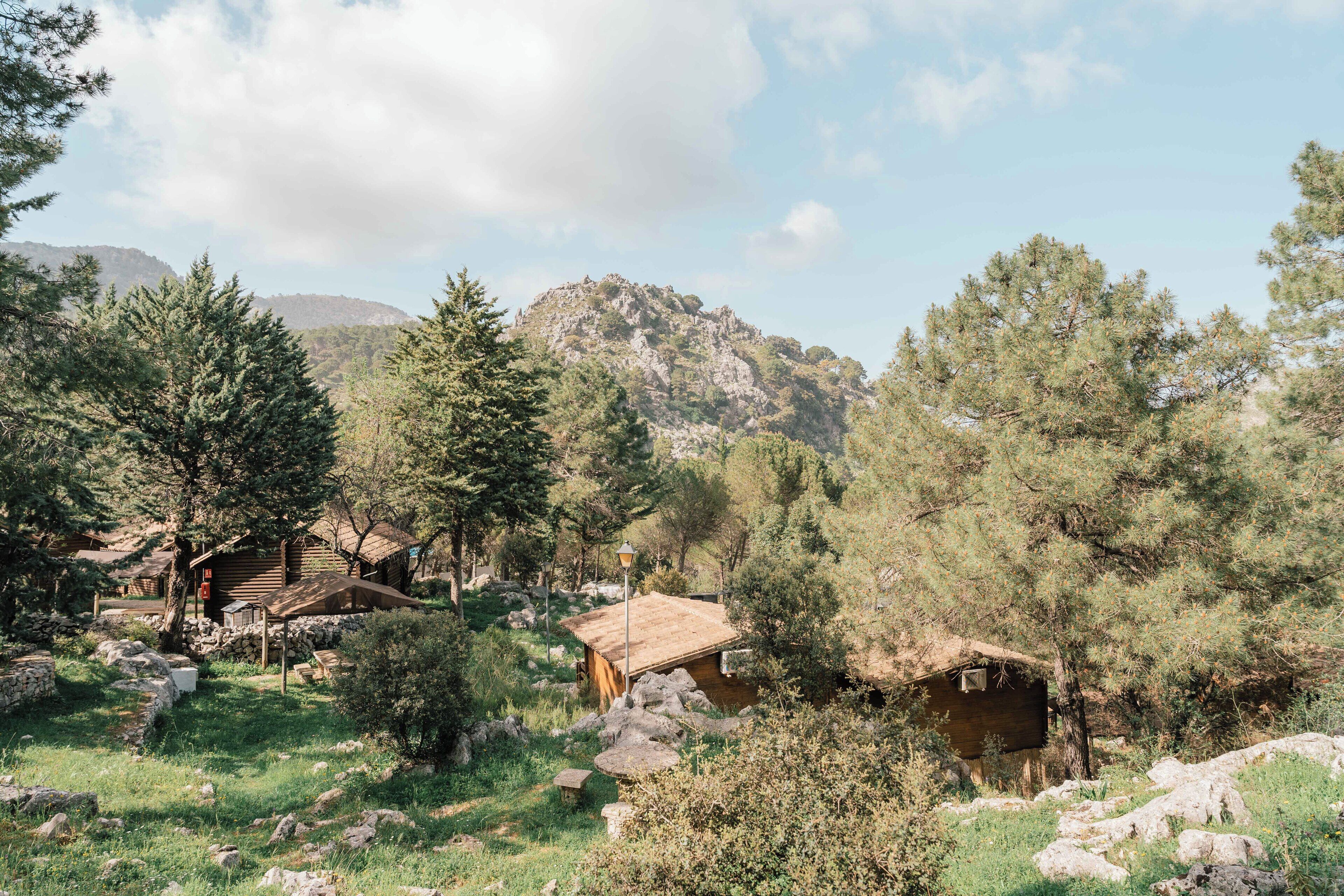 A rustic cabin with a wooden exterior, surrounded by trees and rocky terrain, set against a mountainous backdrop.