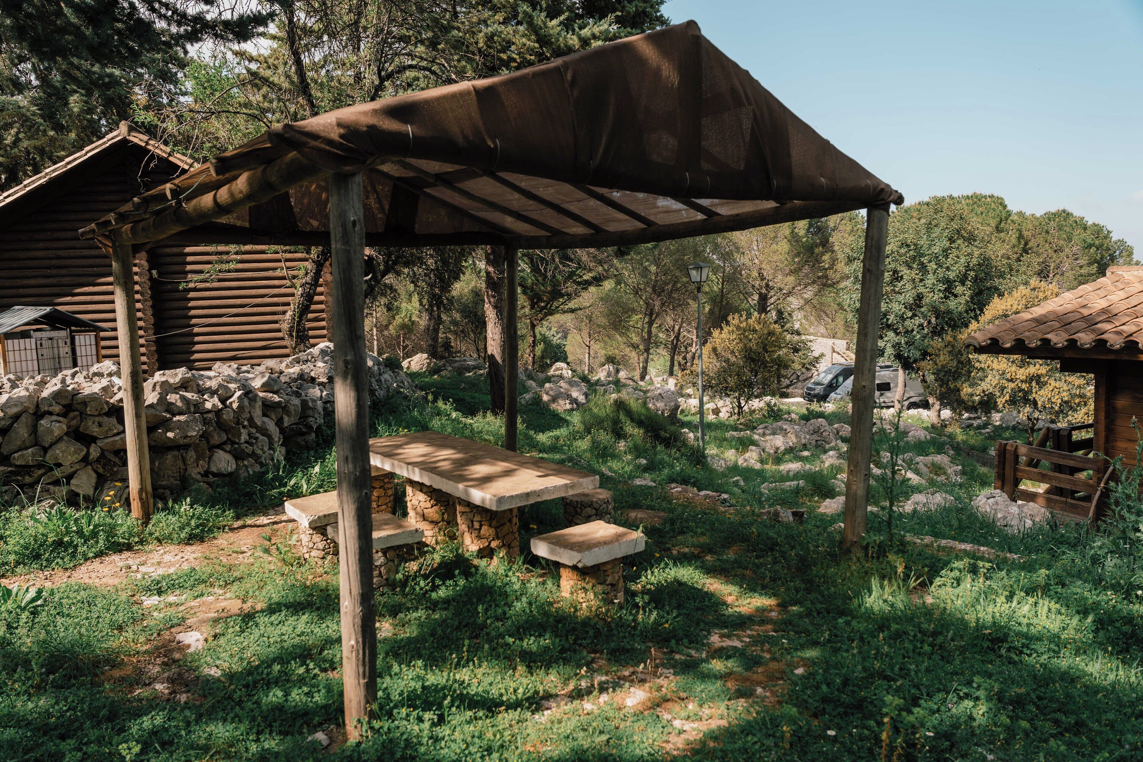 A rustic wooden structure with a stone bench and a covered picnic table, surrounded by greenery and a stone wall.