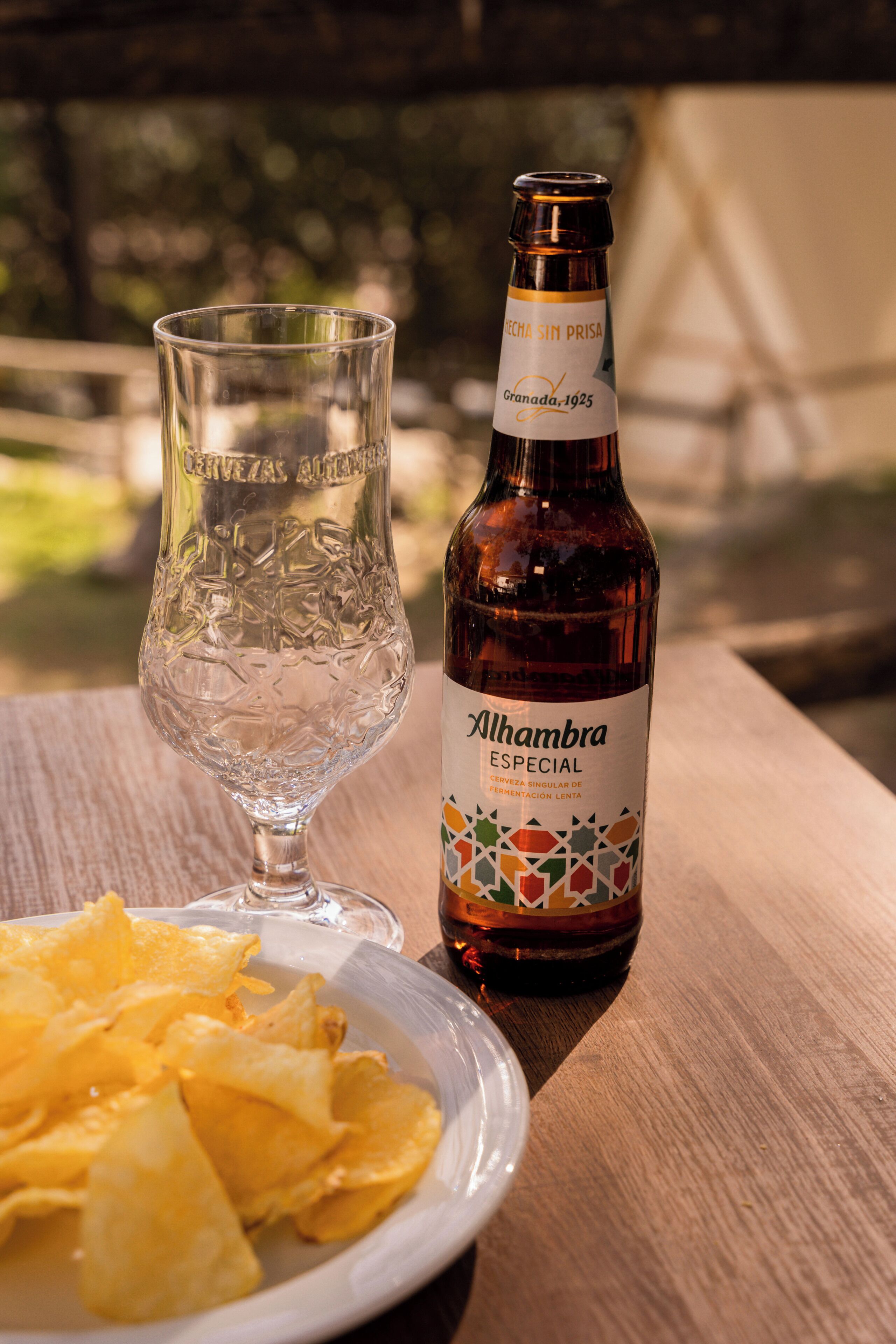 A bottle of Alhambra Especial beer and a glass on a wooden table with a plate of chips.