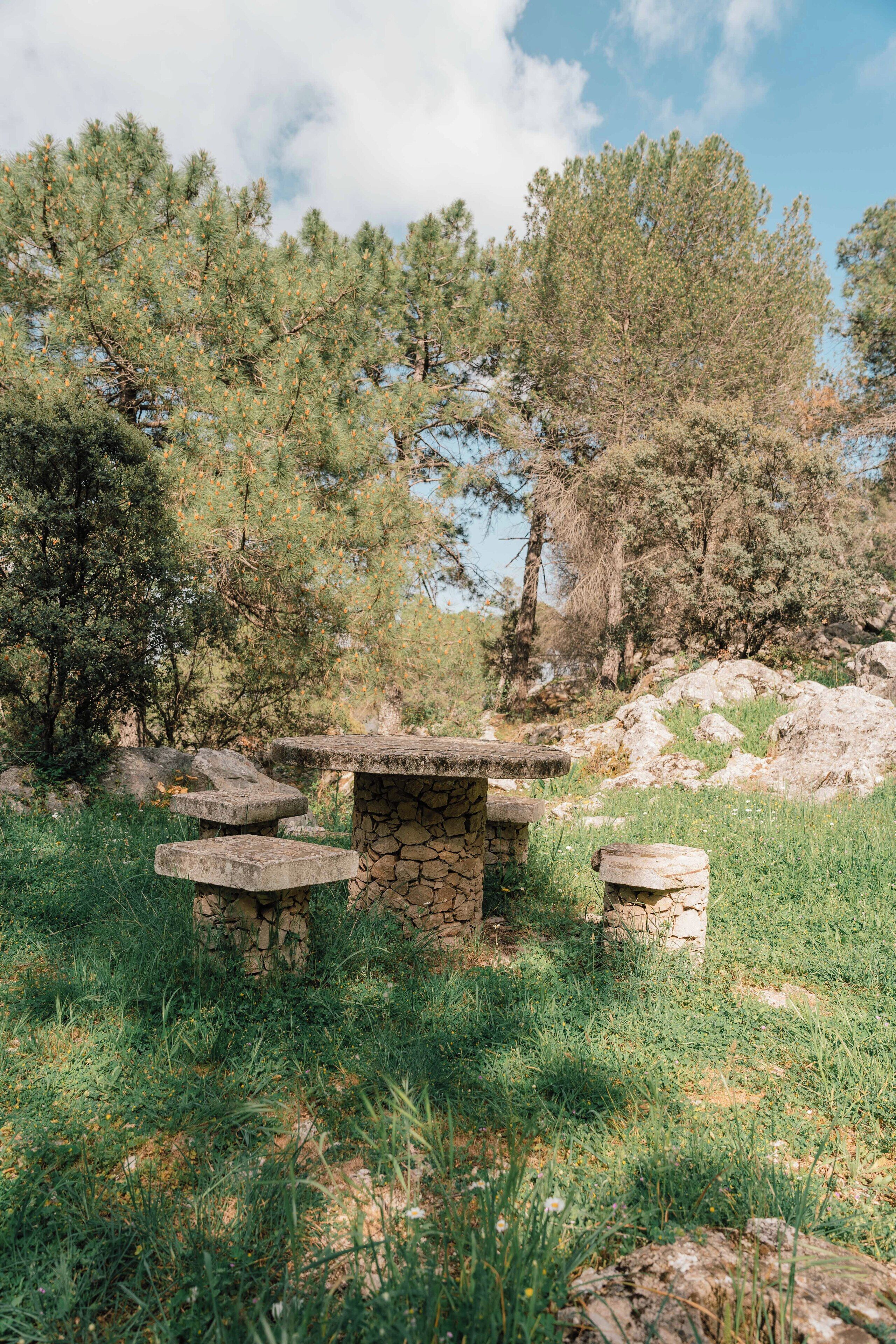 Una estructura de piedra con bancos en un área cubierta de césped, con árboles al fondo.