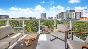Un balcone con una divano e poltrone, un tavolo in vetro con un vassoio di cibo e bevande, e vista su un paesaggio urbano con edifici e gru da costruzione.