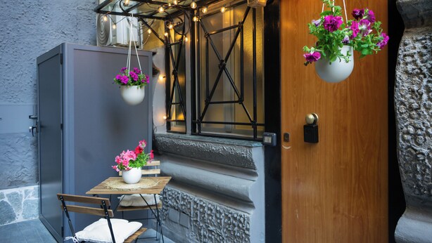 Un petit espace extérieur aménagé avec une table et des chaises, un pot de fleurs suspendu et une porte dont le socle est en pierre.