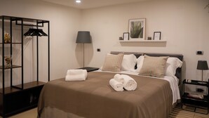 Une chambre d’hôtel avec un grand lit, une table de chevet, une lampe, une étagère avec des objets décoratifs et un porte-vêtements.