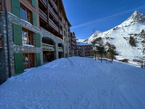 Un edificio de varios pisos con muros de piedra y contraventanas verdes, con una imponente montaña nevada de fondo.
