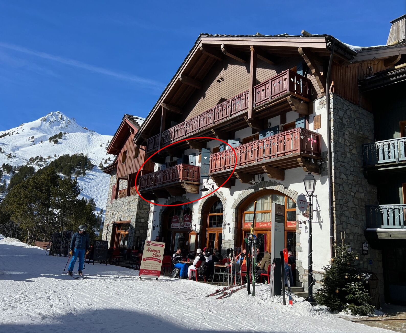 A mountain lodge with wooden balconies and stone walls, a ski slope in the background, and a sign for a restaurant.