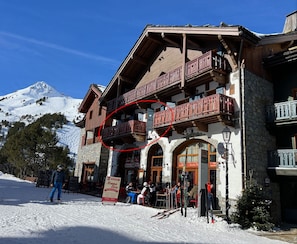 Una cabaña de montaña con balcones de madera y paredes de piedra, una pista de esquí al fondo y un letrero de restaurante.