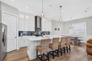 A modern kitchen with white cabinetry, a central island with chairs, and a dining area with a table and chairs. - Luxury Waterfront Townhome | Pool, Boat Slip & Sunset Views (Corpus Christi)