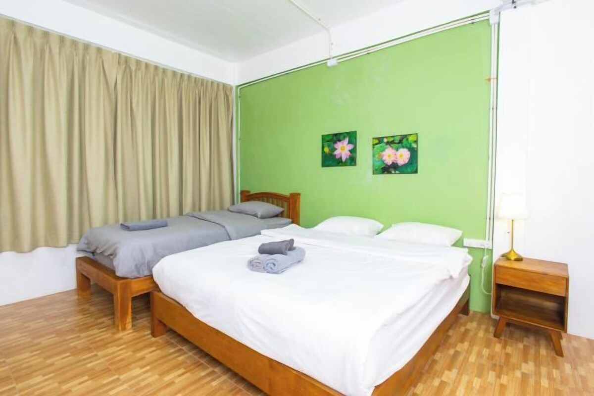 A room with two beds, a wooden floor, a green wall, and a small bedside table with a lamp.