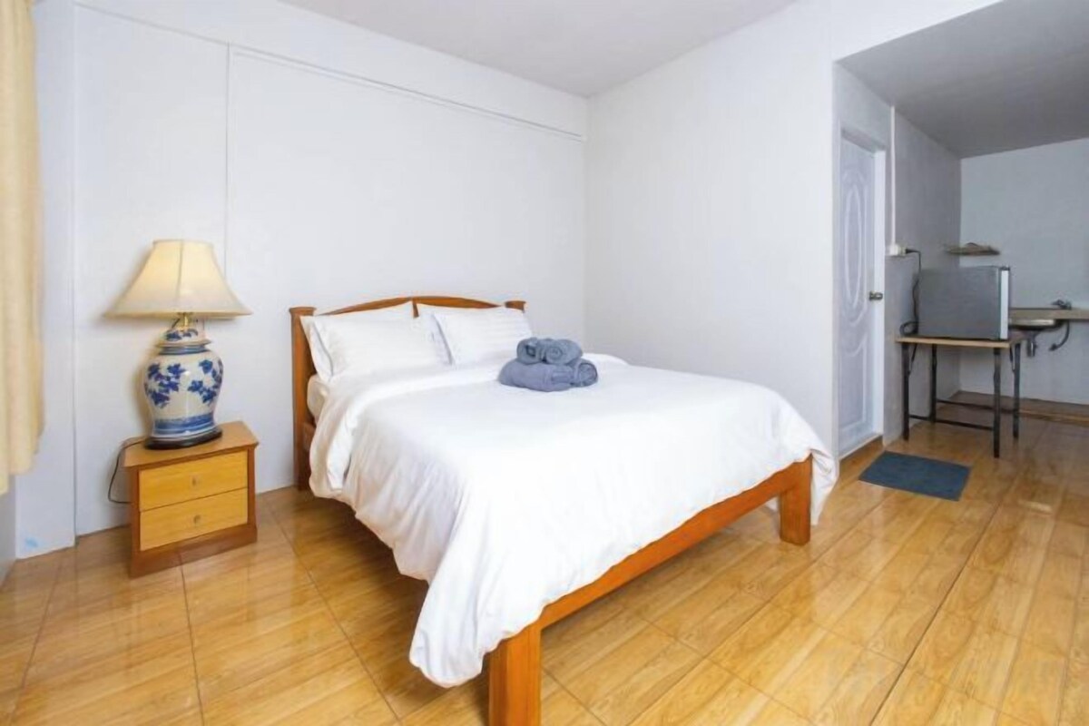 A bedroom with a wooden bed, a bedside table with a lamp, and a desk with a chair.