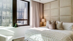 A modern hotel room with a large bed, a sitting area with a chair, a window with curtains, and a city view.