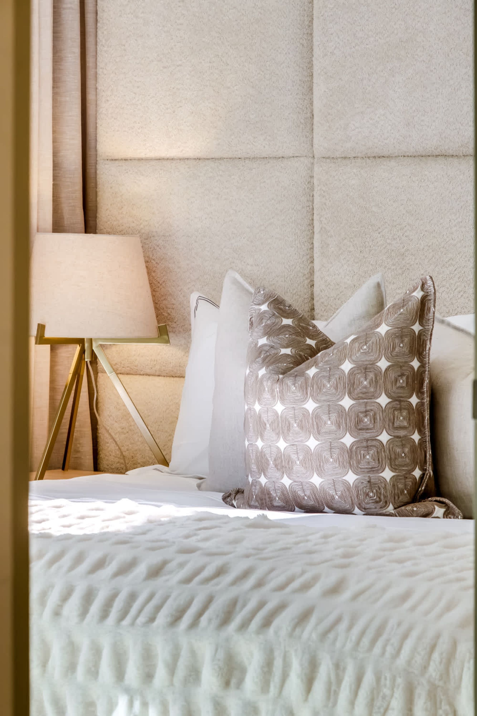 Een net opgemaakt bed met een gebreide witte dekbedovertrek, een gestreept kussen en een gouden nachtkastje met een lamp.