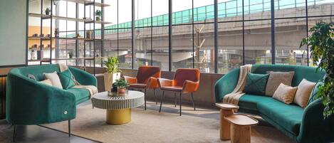 A modern lounge area with two green sofas, a round coffee table, and orange chairs. Large windows offer views of a cityscape and a bridge.
