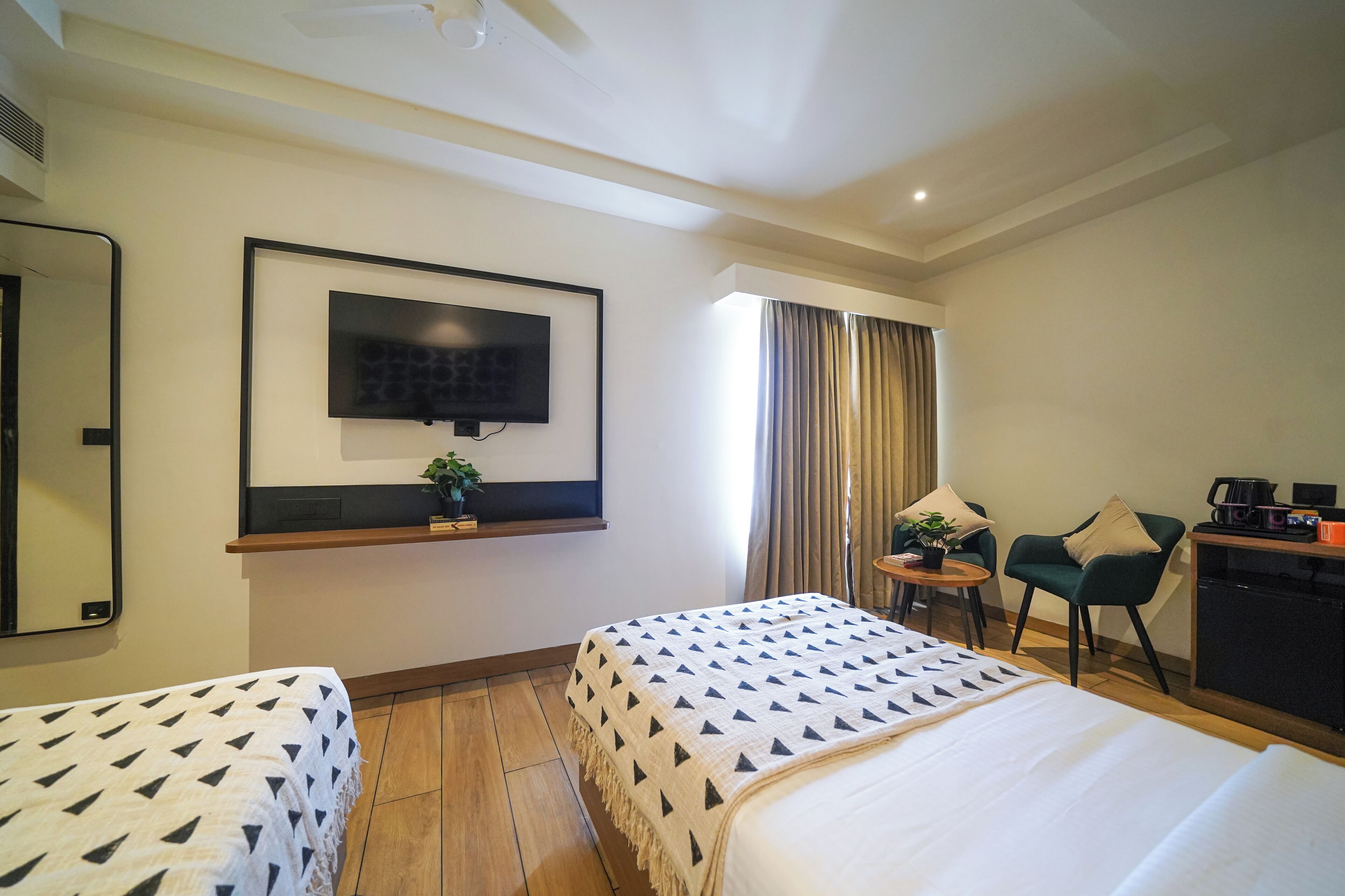 A hotel room with two beds, a TV mounted on the wall, a desk with a coffee maker, and a small table with a plant.