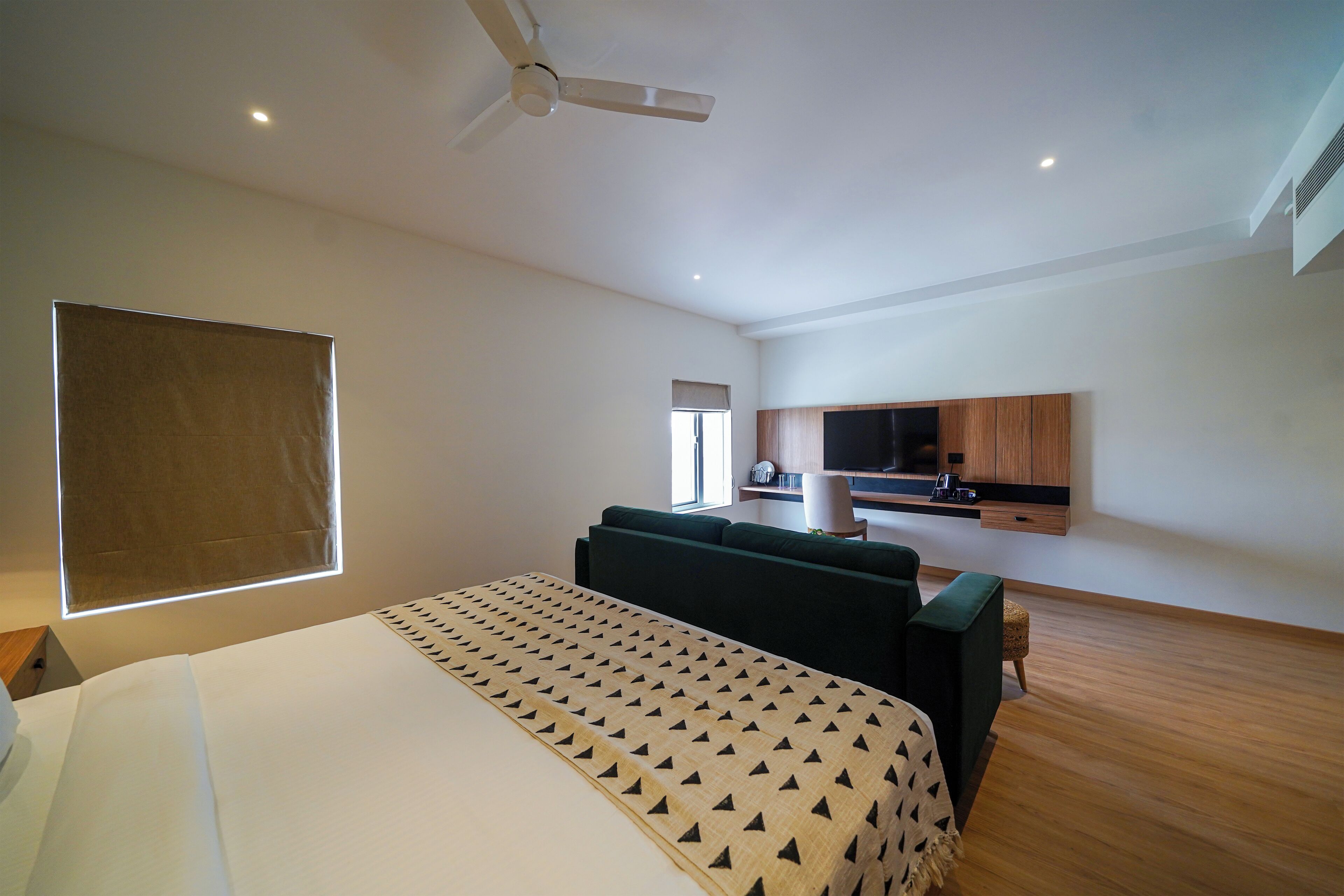 A modern hotel room with a large bed, a green sofa, a small table, a ceiling fan, and a kitchenette in the background.