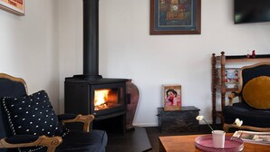 A cozy living room with a black wood-burning stove, a vintage chair, a small table with a vase, and a framed picture on the wall.