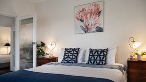 A bedroom with a bed, two bedside tables with lamps, a door with frosted glass, and a wall-mounted painting of flowers.