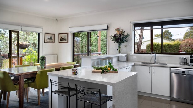 A modern kitchen with a central island, dining area with chairs, and large windows offering views of greenery.