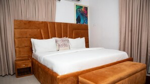 A bedroom with a large bed, a wooden nightstand, a bench, and a colorful abstract painting on the wall. - Micasa Haven (Lekki)