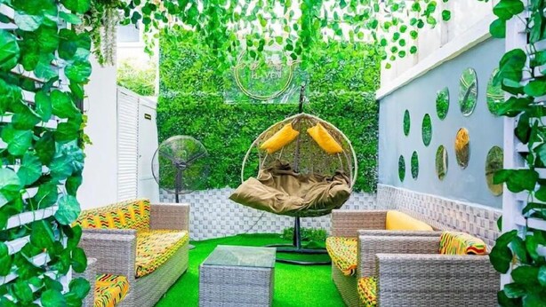 A cozy outdoor seating area with wicker furniture and a hanging chair, surrounded by lush greenery and a white wall with decorative leaf patterns. - Micasa Haven (Lekki)