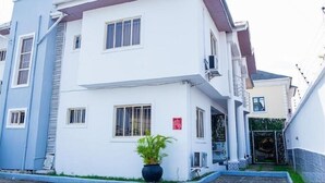 A two-story building with a white facade, multiple windows, and a small garden area with a potted plant. - Micasa Haven (Lekki)