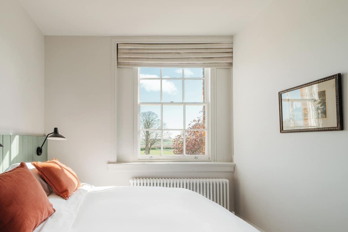 A bedroom with a large window, a radiator, a bed with white bedding and two orange pillows, and a framed picture on the wall.