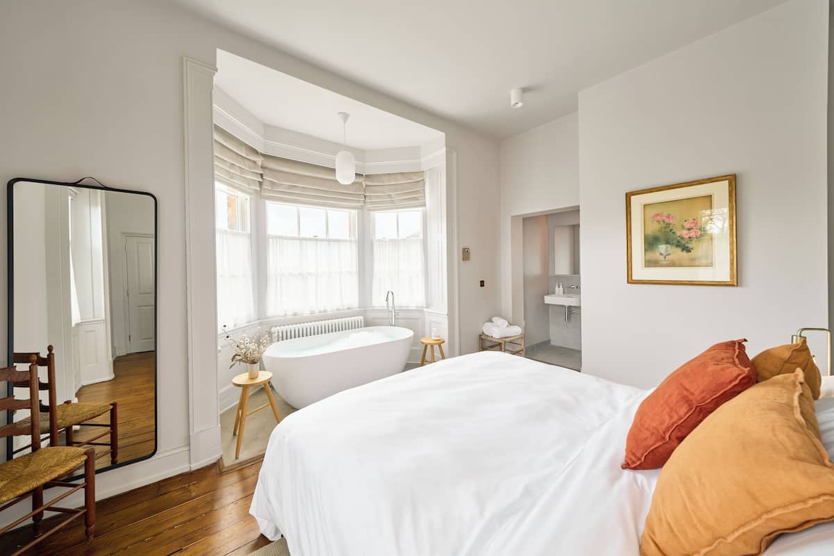 A spacious bedroom with a large bed, a sitting area with a chair, a bathtub, and a mirror.