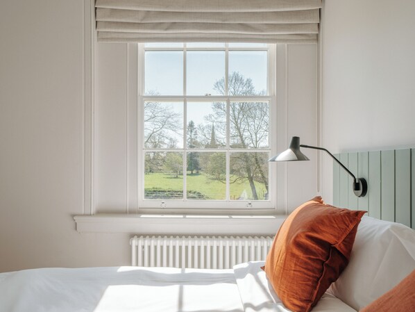 A bedroom with a large window, a radiator, a bed with white bedding and an orange pillow, and a black desk lamp.