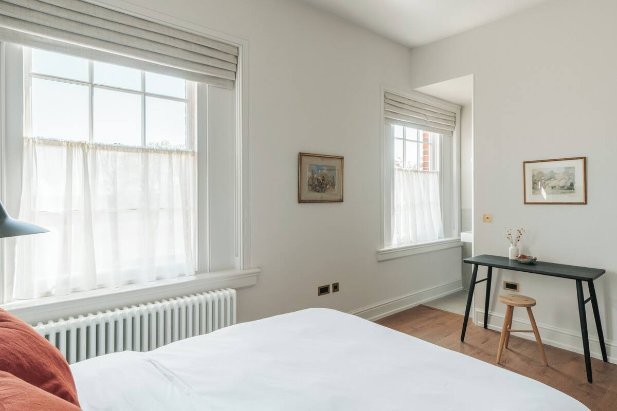 A bedroom with a large bed, two windows with blinds, a radiator, a desk with a chair, and two framed pictures on the wall.